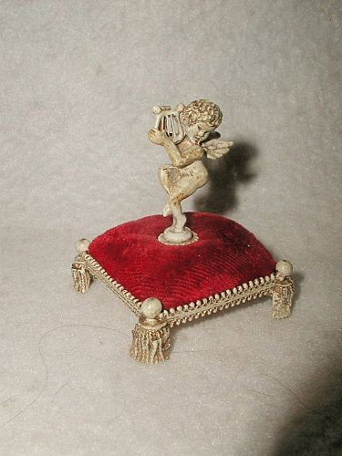 Cherub Pin Cushion France Velvet Metal Tasseled Base Ea: Unusual French early 1900's cherub pin cushion in red velvet atop an antique cream metal stool with metal trim on all sides standing on 4 tasseled feet. Antique cream and gilt cherub in center holding