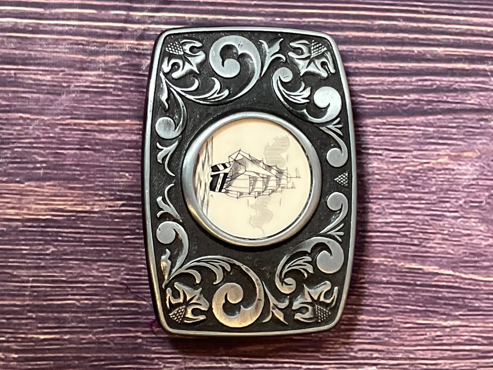 Vintage Scrimshaw-Style Sailing Ship Belt Buckle – Black Metal – Nautical Maritime Design: vintage nautical-themed belt buckle featuring a scrimshaw-style image of a classic sailing ship set in a round inset. The buckle is made of black-finished metal with an ornate scroll border and floral