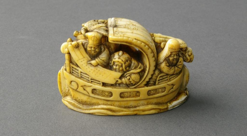 JAPANESE CARVED IVORY NETSUKE - GODS OF FORTUNE: depicting seven figures, likely the "Seven Gods of Good Fortune", crowded on the deck of a dragon-form sailing ship, signed, 2in. SMALL CHIP ON BASE HAS OLD REPAIR NO INTERNATIONAL SHIPPING.