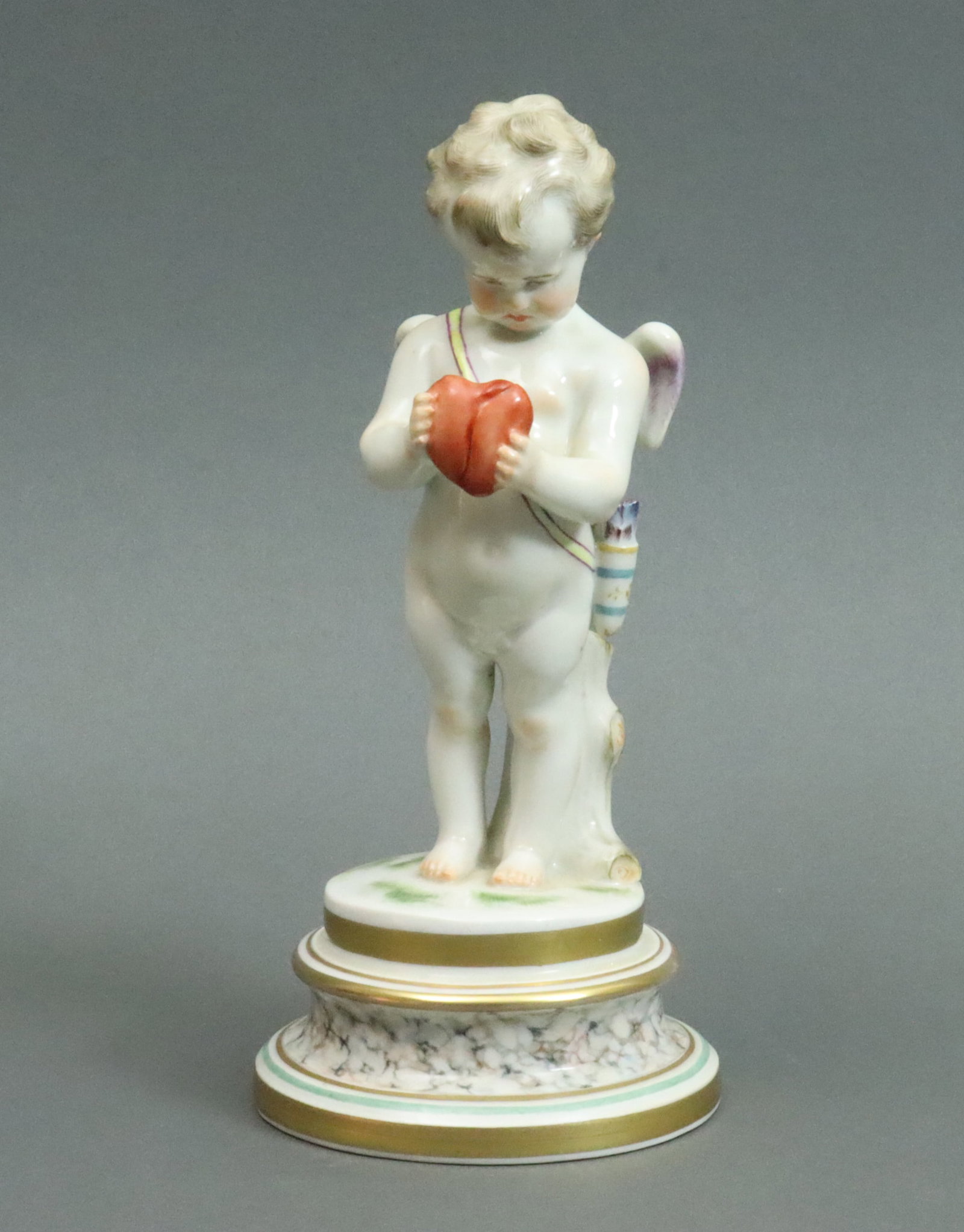 MEISSEN PORCELAIN BROKEN HEART FIGURINE: depicting a winged cupid wearing a quiver, leaning on a tree stump, holding a broken heart, raised on a marbleized plinth, blue underglaze crossed swords mark, UV clean, no condition issues, 8.25in., 