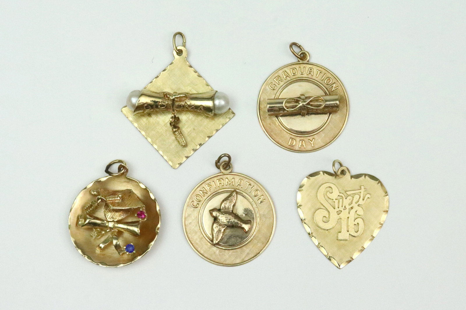 (5) 14K CHARMS: including a double pearl diploma with dangling tassel, a Sweet Sixteen heart with inscription, a gem-set graduation cap with articulated tassel dated 82, a Graduation Day diploma dated 74, and a Confi