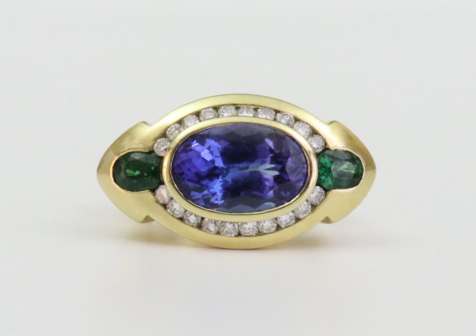 18K TANZANITE, GARNET & DIAMOND RING: bezel-set with a central oval (8x12mm) tanzanite within a surround of 20 round channel-set natural diamonds approx. 0.40ctw, and flanked by 2 (5x4mm) green garnets (likely tsavorite), overall ring
