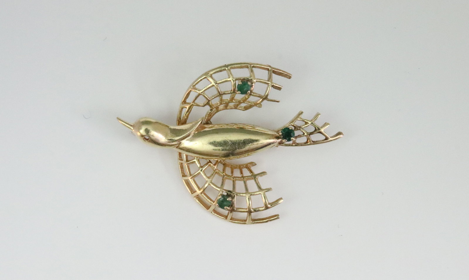 14K STYLIZED BIRD PIN WITH EMERALD ACCENTS: in openwork wing and tail design, set with 3 round emeralds approx. 0.20ctw, in yellow gold, stamped 14K, slight bend to pin stem, missing eye gem, 1.6x1.2in., 5.8gms., Opening Bid $300