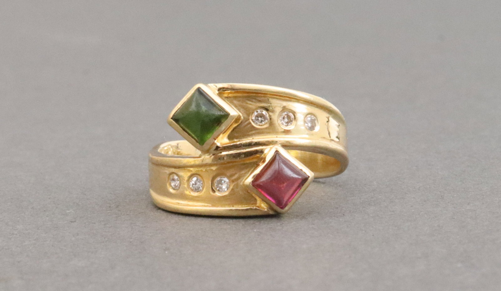 18K MULTI GEMSTONE BYPASS RING: comprising 6 round flush-set natural melee diamonds, and 2 square-cut colored gemstone terminals, likely emerald and amethyst cabochons each 0.35ct (stone scuffs), height 12mm, in yellow gold, stamped