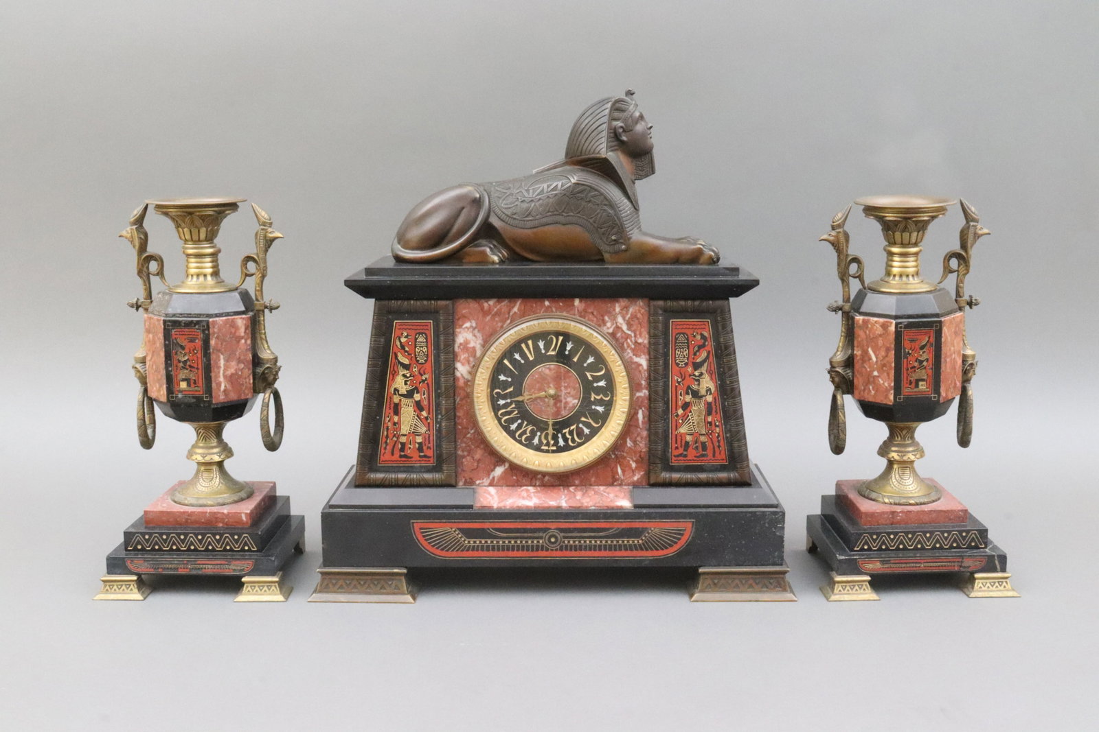ETIENNE MAXANT EQYPTIAN REVIVAL MANTLE GARNITURE: c.1880, in bronze, slate and rouge marble, the Sphinx in recumbent pose above the clock case, the case decorated in matching panels depicting a divinity figure and hieroglyphic symbols, the clock