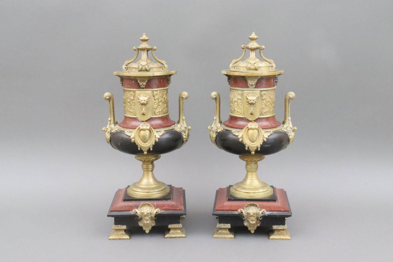 PAIR GILT BRONZE NEOCLASSICAL-STYLE CAMPANA URNS: in rouge marble and slate, with scrolling handles, lion masks and scrollwork relief bands, above a square tiered plinth centered with a Bacchus mask within a shell cartouche, removable urn lids, one