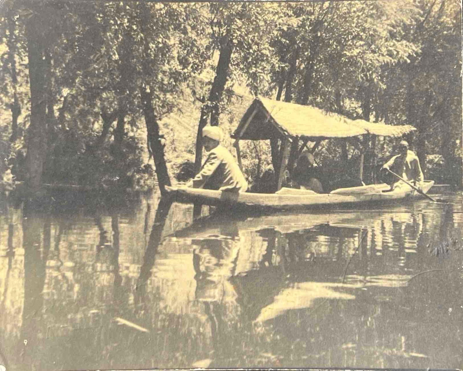 JOURNEY THROUGH THE QUIET WATERWAY: JOURNEY THROUGH THE QUIET WATERWAY THIS SEPIA-TONED PHOTOGRAPH CAPTURES A PEACEFUL SCENE OF THREE PEOPLE TRAVELING ALONG A CALM, TREE-LINED RIVER IN A NARROW WOODEN BOAT. THE BOAT FEATURES A SMALL