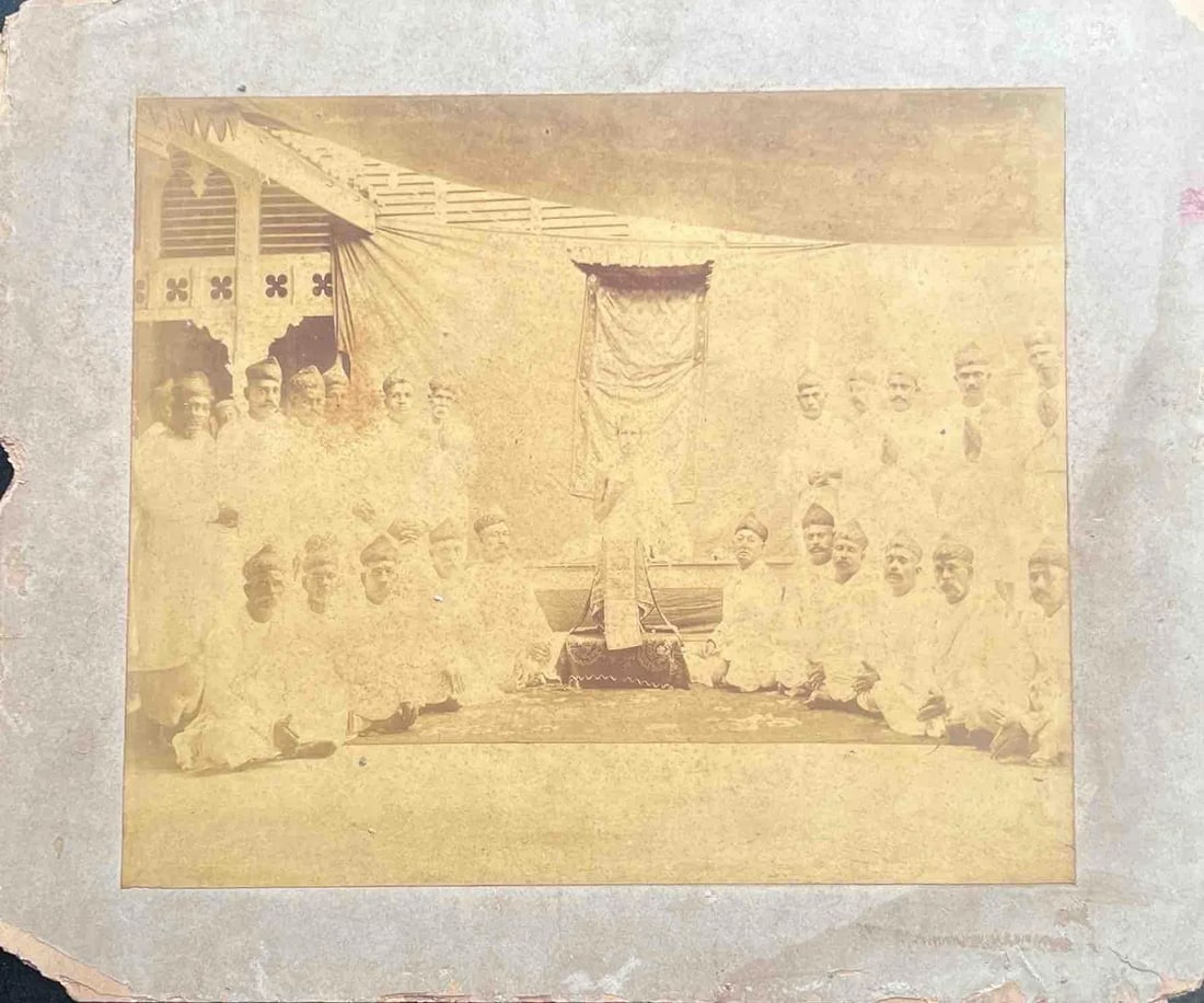 VINTAGE GROUP PORTRAIT OF A CEREMONIAL GATHERING: VINTAGE GROUP PORTRAIT OF A CEREMONIAL GATHERINGAN OLD, FADED SEPIA-TONED PHOTOGRAPH SHOWING A LARGE GROUP OF MEN DRESSED IN TRADITIONAL ATTIRE, SEATED AND STANDING AROUND A CENTRAL FIGURE UNDER A DEC
