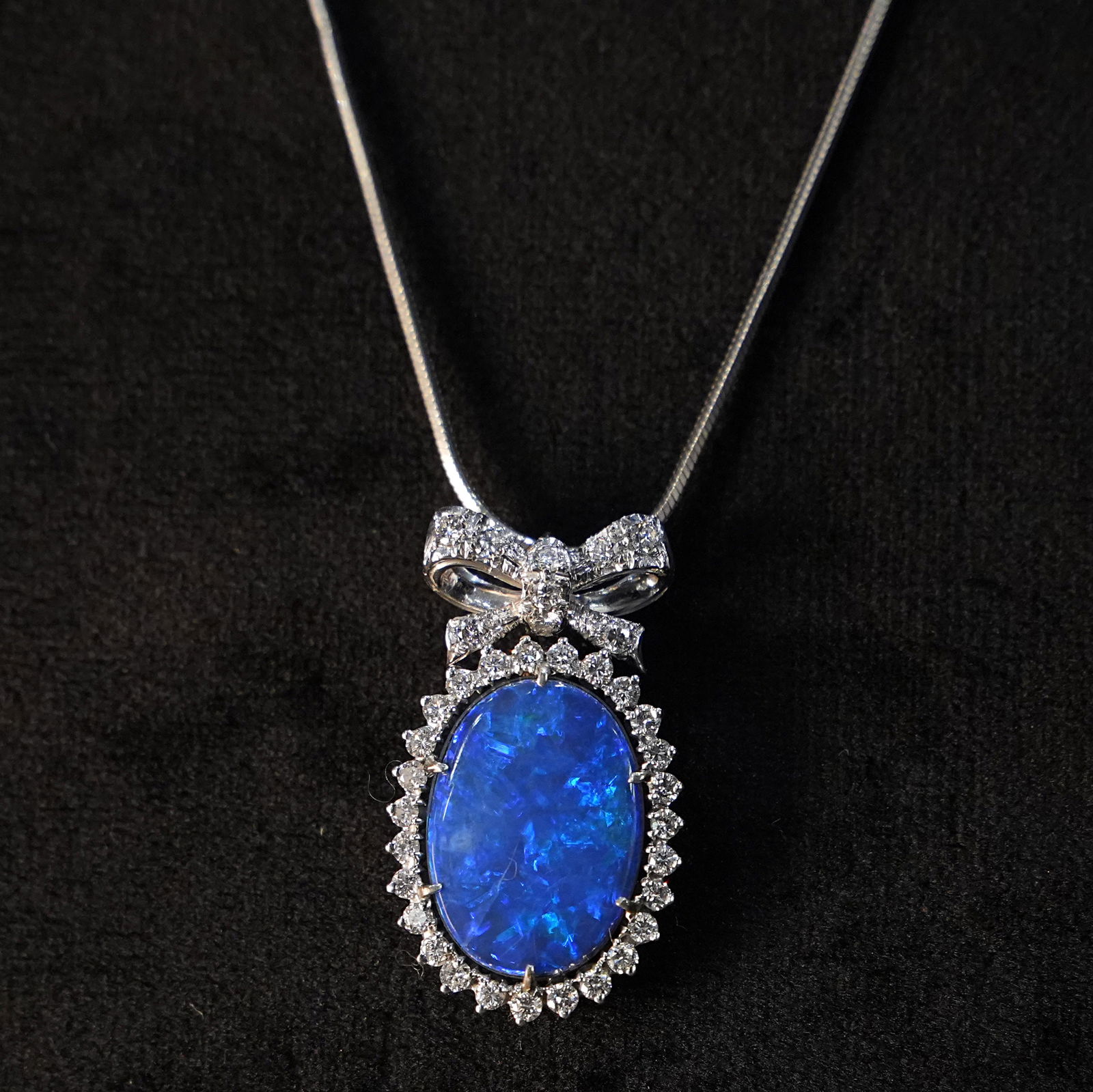 18K White Gold Pendant with Blue Opal Surrounded by Diamonds and Gold Bow with Diamonds 4.46g: Graceful pendant crafted in 18K white gold, featuring a luminous blue opal encircled by sparkling diamonds, surmounted by a graceful bow also adorned with diamonds. The elegant combination of the opal