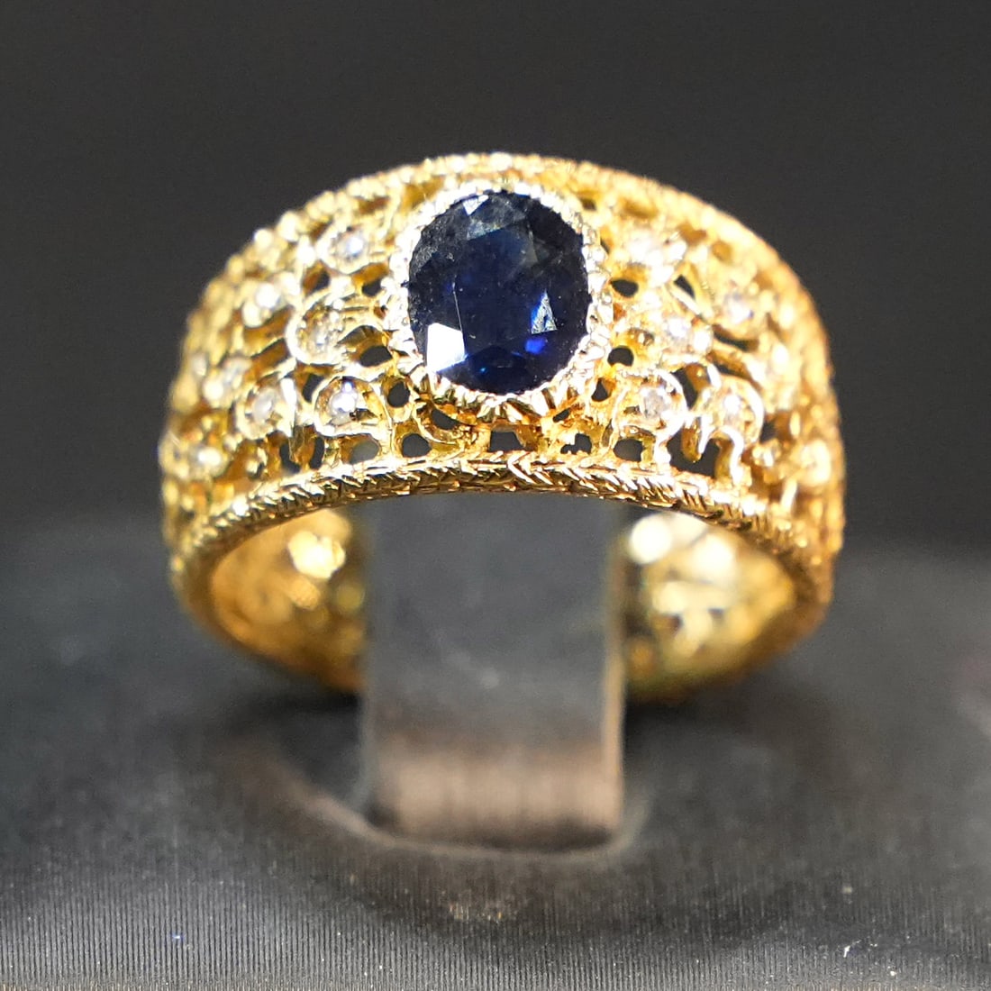 Delicate 18K Yellow Gold Filigree Band Ring with Central Sapphire and full of 0.3ct Diamonds, 4.99gr: Delicate 18K yellow gold ring featuring a wide filigree band with delicate lace-like motifs, enhancing a central sapphire for a luminous focal point. The airy yet sculpted structure creates an etherea