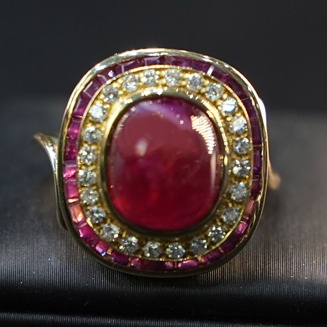 18K Yellow Gold Ring with Cabochon Ruby, Ruby Halo (6.00ct total), 0.30ct Diamonds, 9.26gr: An elegant ring fashioned in 18K yellow gold, showcasing a large central cabochon ruby with a smooth, domed profile. The centerpiece is encircled by a bright diamond surround and an additional outer c