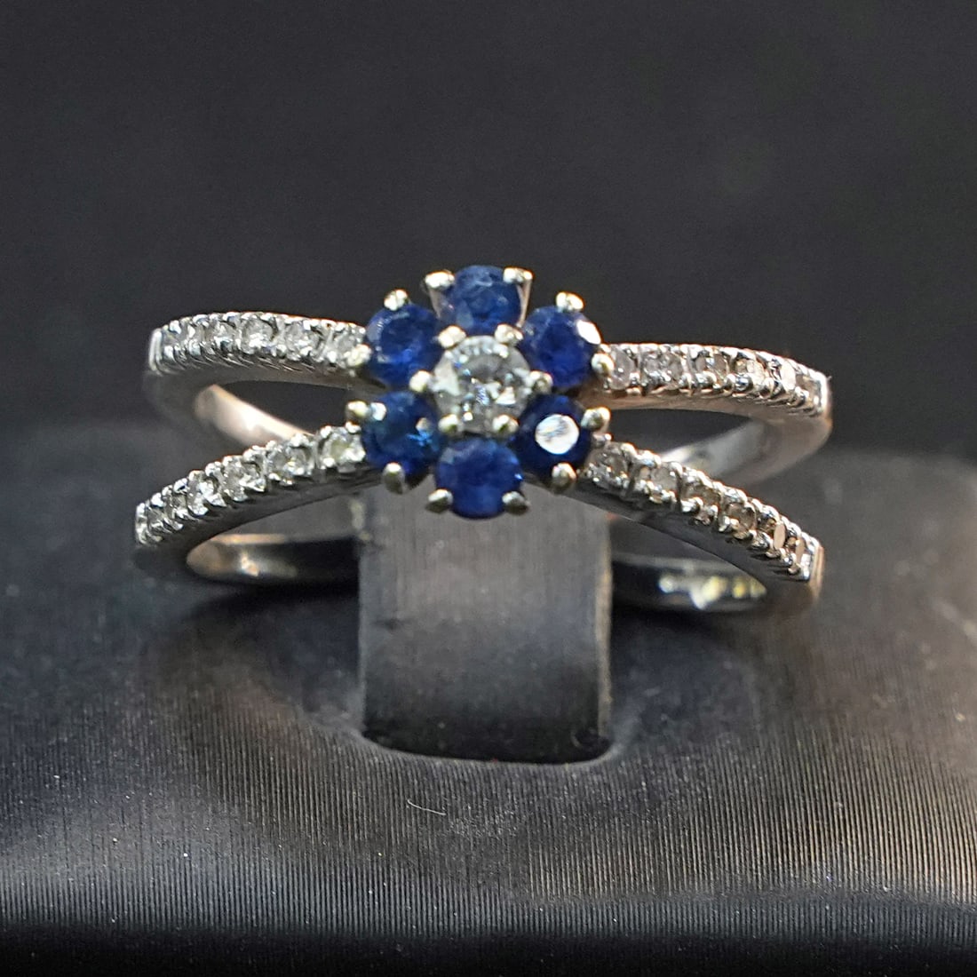 Original 18kt White Gold Ring with Sapphire Halo, Central Diamond and Side Diamonds, 6.32 gr: Original 18kt white gold ring designed with a refined floral-inspired sapphire halo surrounding a central diamond, complemented by additional diamonds set along the sides, creating depth and elegant m