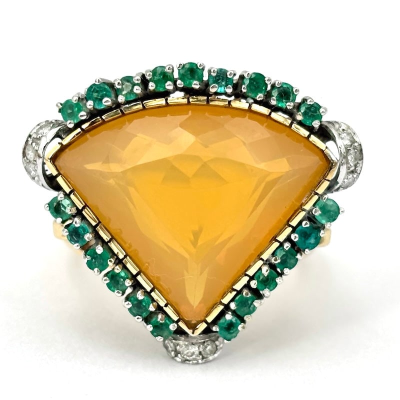Triangular ring in fire opal style, emeralds, diamonds and gold; 12.66 gr.: Spectacular and highly original maxi triangle ring rounded at the base in style, in 18 kt gold with perimeter emeralds, rhinestones at the apexes and a diamond-cut fire opal as the eccentric central s