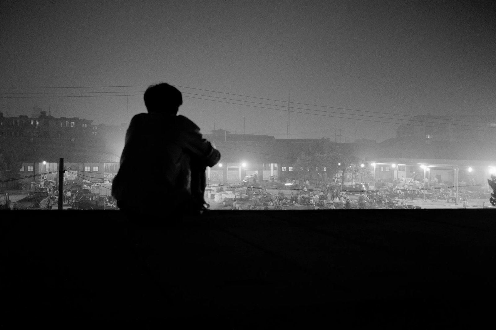 The Bustle Belongs to Others: Series: Migrant Workers Series Caption: A migrant worker sits on a bridge, gazing toward the bustling market in the distance. Print Date: 2018 Medium: Cotton fine art paper, archival giclee print