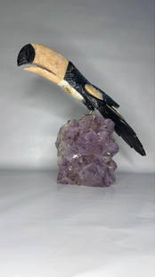 Brazilian Gemstone Toucan Sculpture on Amethyst Cluster: A striking Brazilian toucan sculpture, expertly carved from semi-precious gemstones, perched on a radiant cluster of natural amethyst crystals. The bird&#8217;s bold silhouette and lifelike detail giv