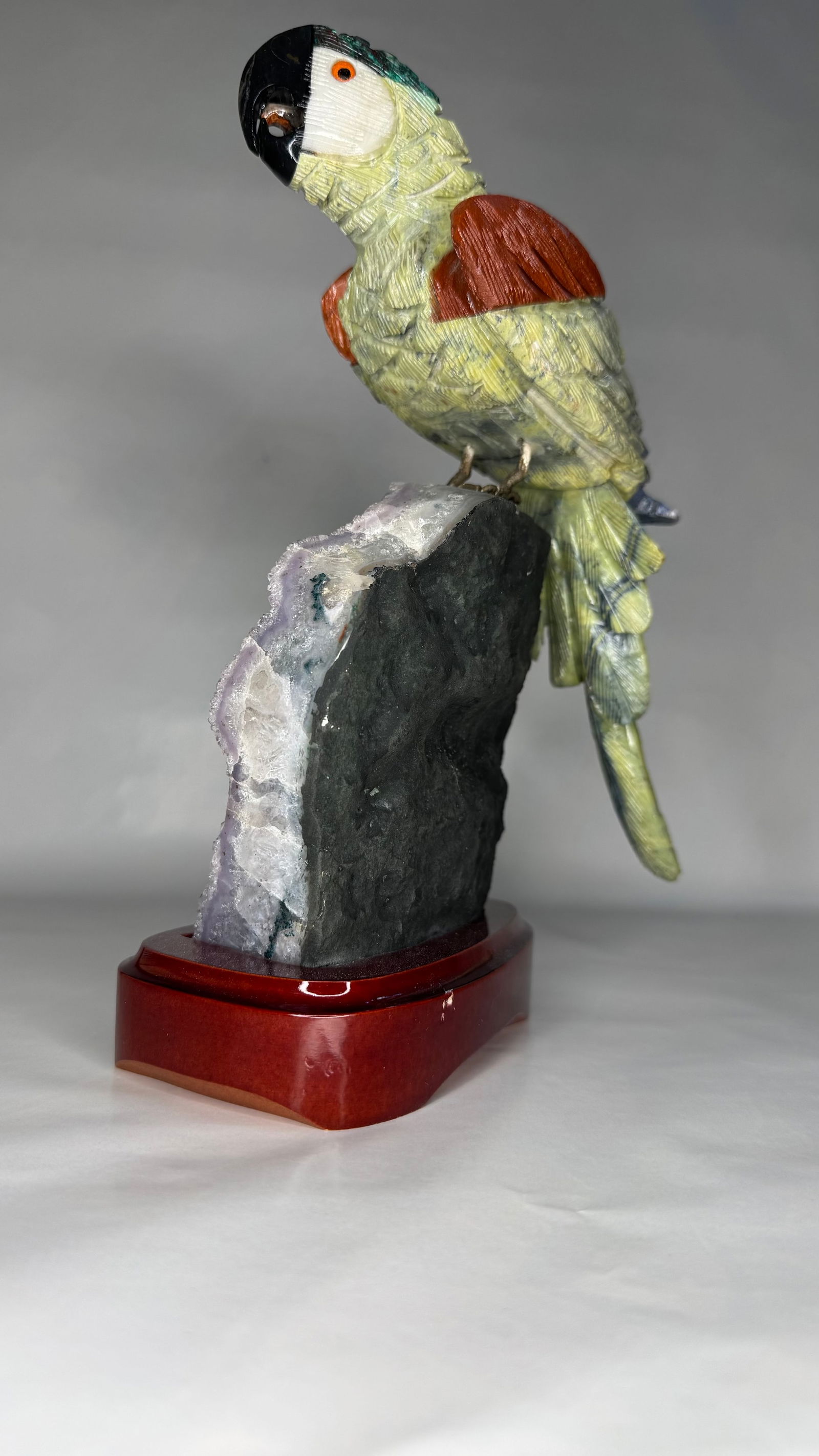 Brazilian Gemstone Parrot Sculpture on Amethyst Cluster with Wooden Stand: An exquisite hand-carved parrot sculpture composed of assorted semi-precious Brazilian gemstones, intricately assembled to capture the vibrant character of this tropical bird. The body, wings, and bea