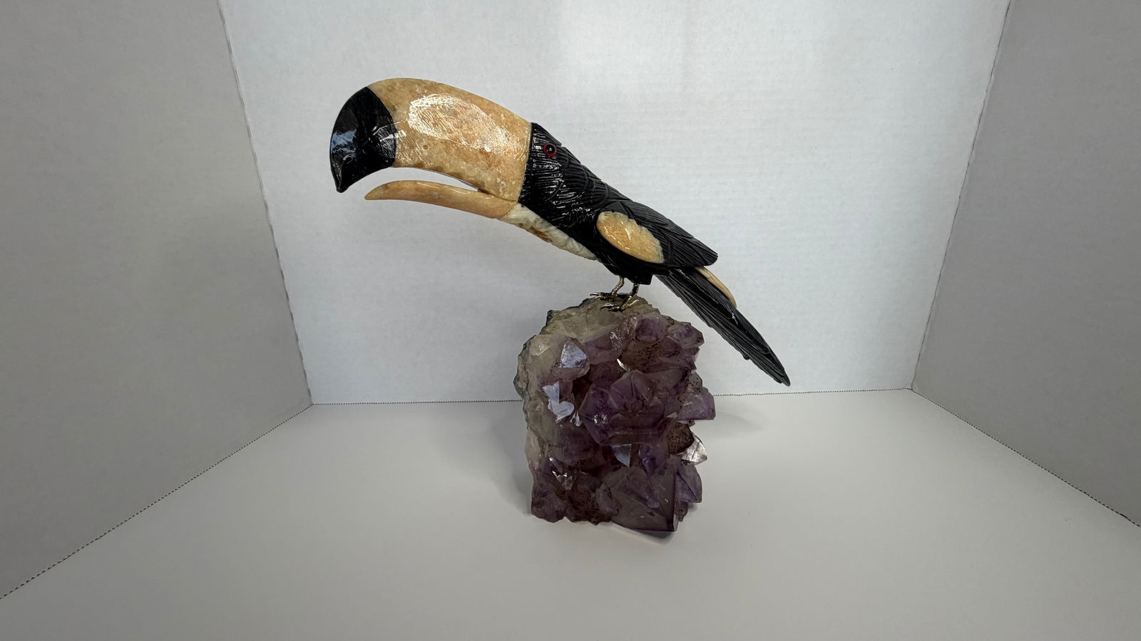 Brazilian Gemstone Toucan Sculpture on Amethyst Cluster: A striking Brazilian toucan sculpture, expertly carved from semi-precious gemstones, perched on a radiant cluster of natural amethyst crystals. The bird's bold silhouette and lifelike detail give it a