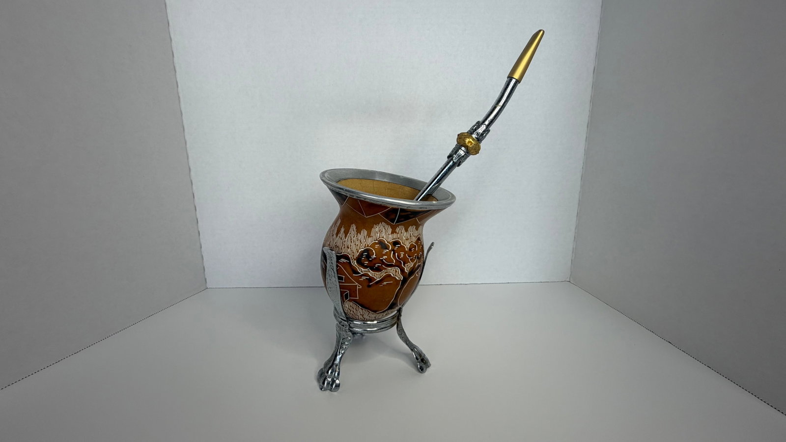 Traditional Brazilian Mate Gourd with Bombilla and Ornate Stand: A finely handcrafted traditional mate gourd set from Brazil, comprising a natural gourd cup with engraved detailing, an ornate aluminum stand, and a classic metal bombilla for sipping. The gourd