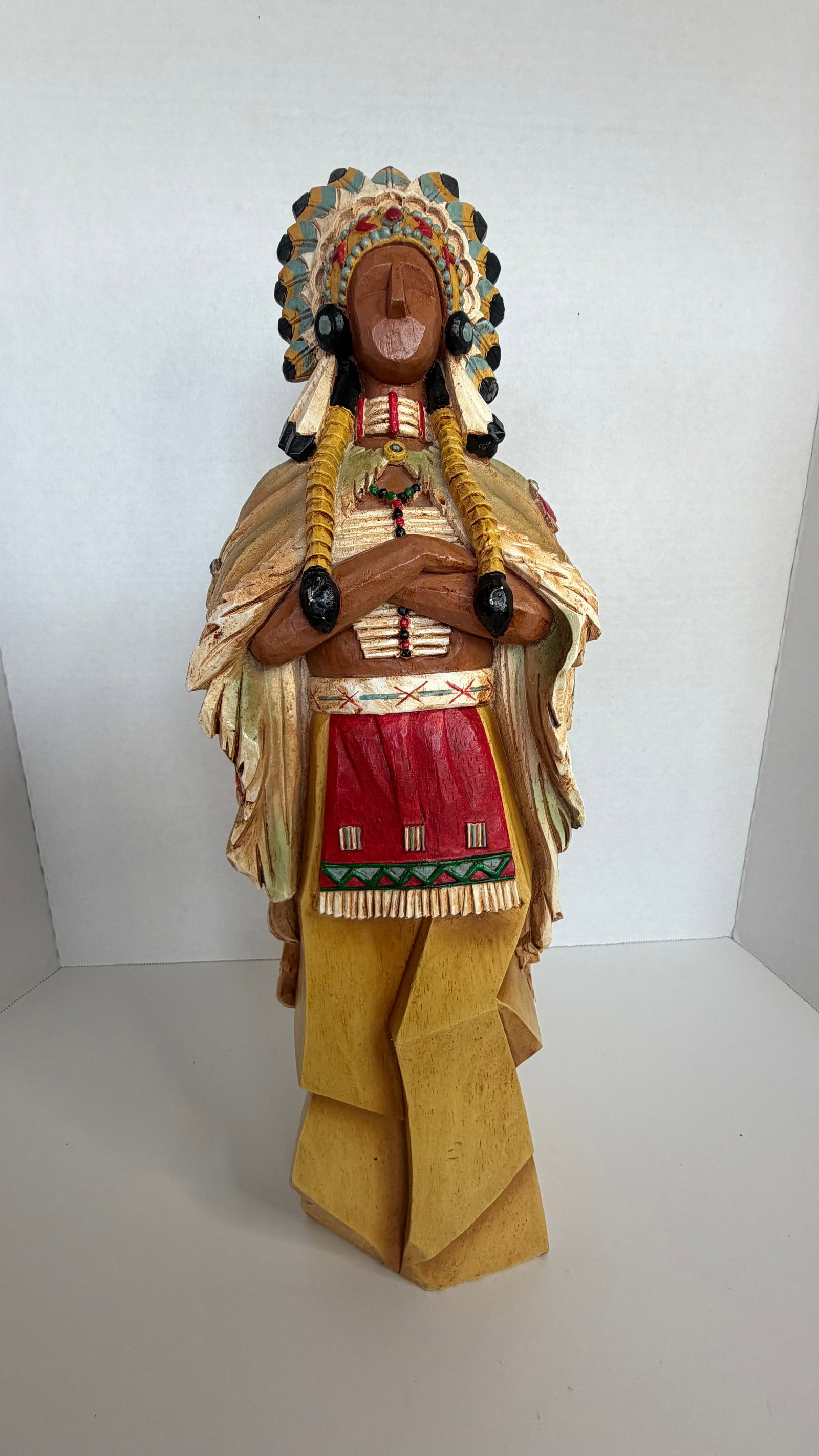 Hand-Carved Wooden Sculpture Native American Figure with Stylized Headdress and Geometric Form: A powerful and visually compelling hand-carved wooden sculpture, depicting a Native American figure, likely a chief or elder, in a dignified, upright stance with arms folded across the chest and head