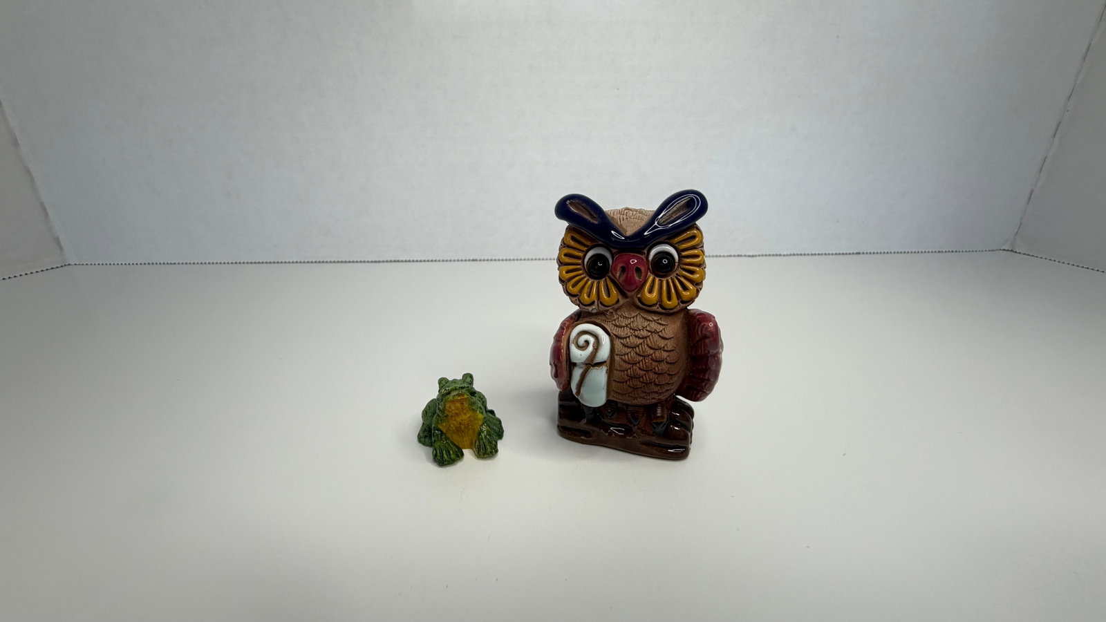 Hand-Painted Ceramic Figurine Set: Owl with Scroll & Frog Companion: A charming hand-painted ceramic figurine duo featuring a whimsical owl clutching a scroll, paired with a playful green frog companion. The owl is rendered with expressive eyes, finely textured feather