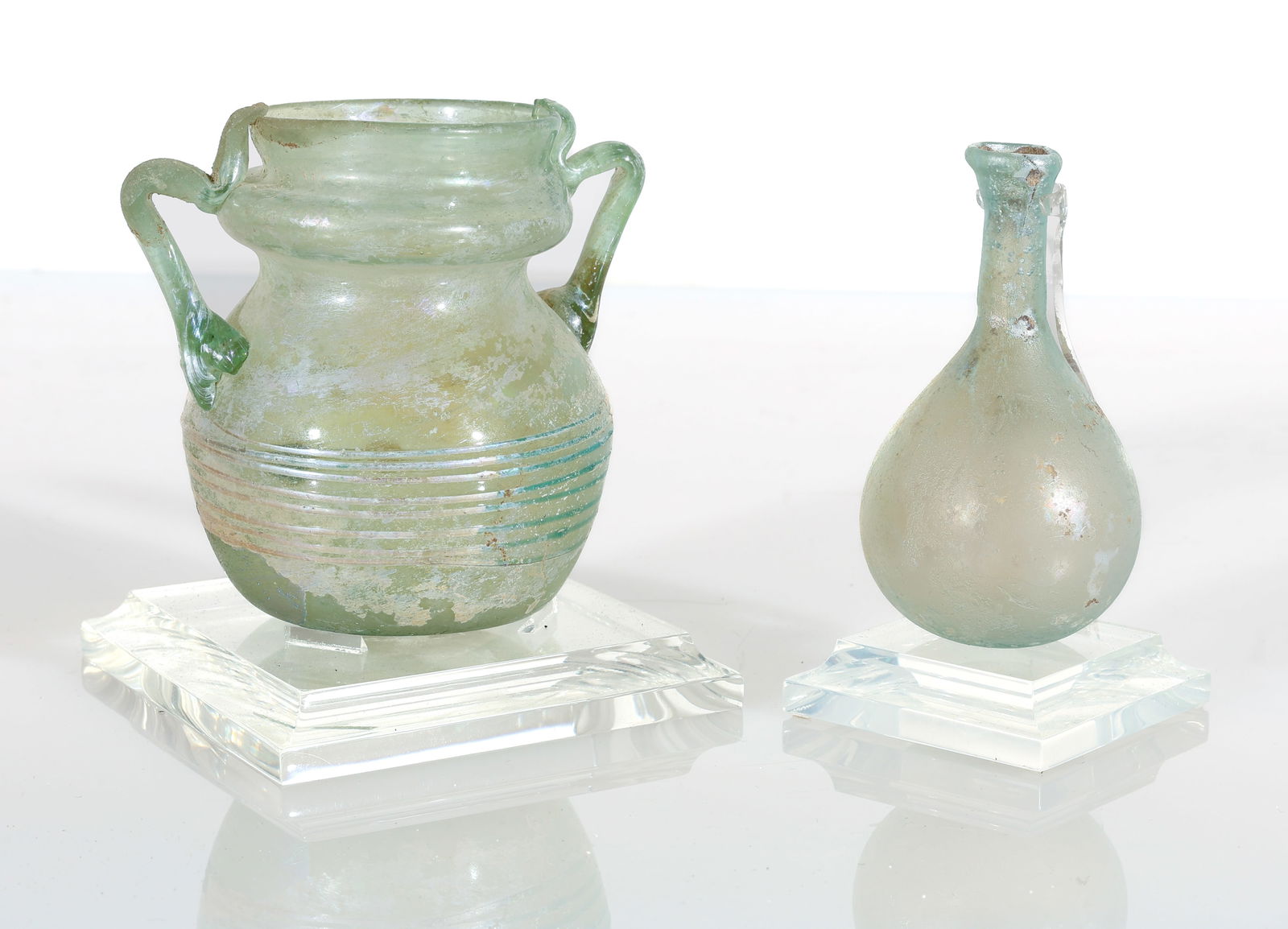 (2) Ancient Roman Glass Cosmetic Bottles: (2) Ancient Roman Glass Cosmetic Bottles, each with display stand. Sizes: 3.25"H (11 cm) and 3.25" (8 cm).