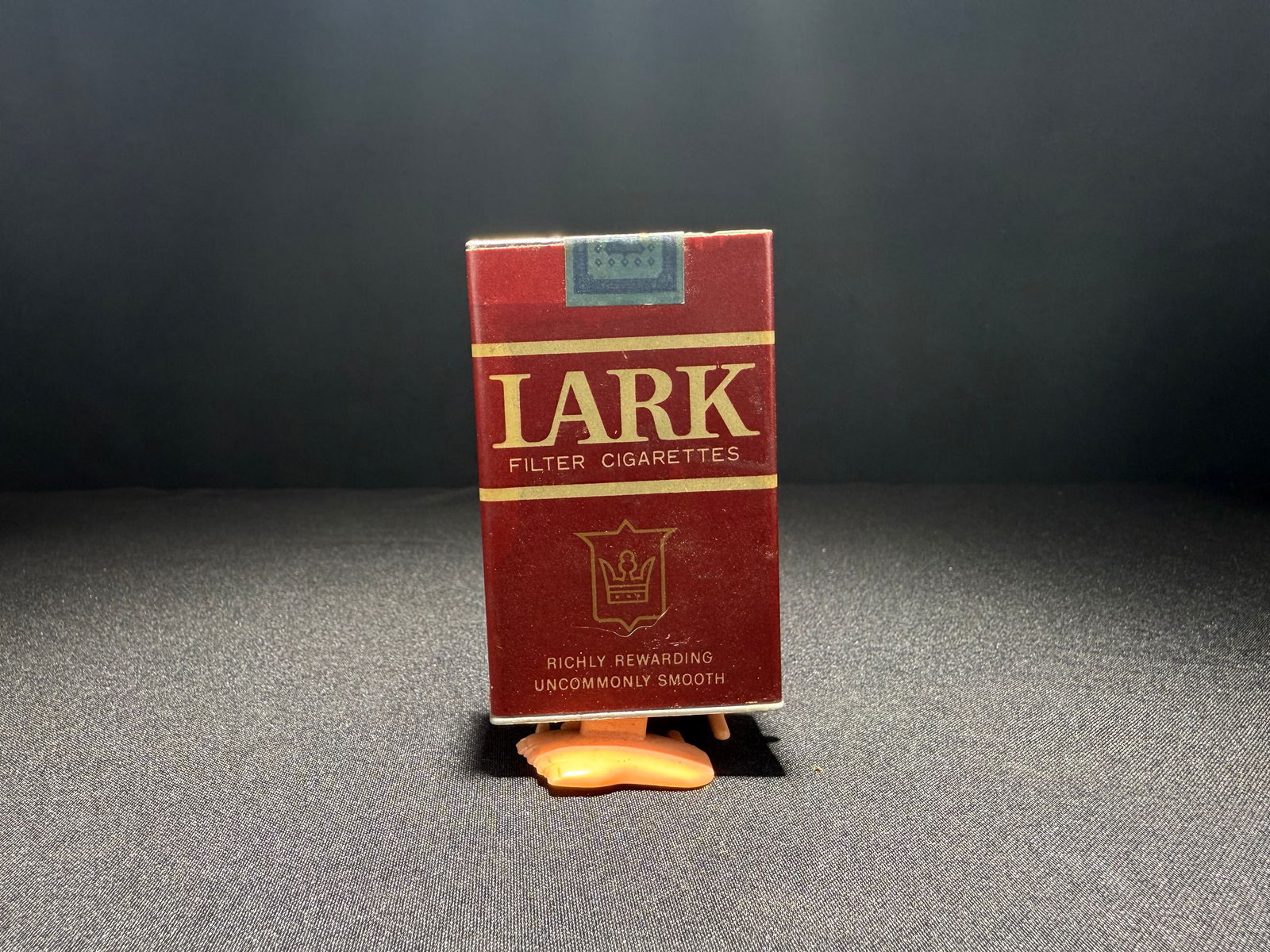 Lark Cigarettes Walking Pack: Novelty promotional item featuring a mechanical walking Lark cigarette pack with flesh-colored plastic feet. The maroon and gold pack is detailed with period-accurate Lark branding, including the crow