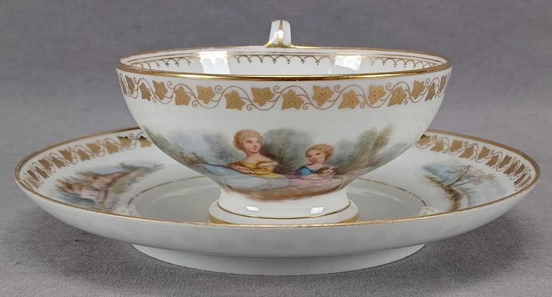 Sevres Hand Painted Watteau Scene Tea Cup & Saucer: Antique footed porcelain tea cup and saucer. Decorated with a hand painted Watteau scene with two ladies on the cup. The saucer has hand painted landscape scenes on either side. Gold ivy leaf band aro