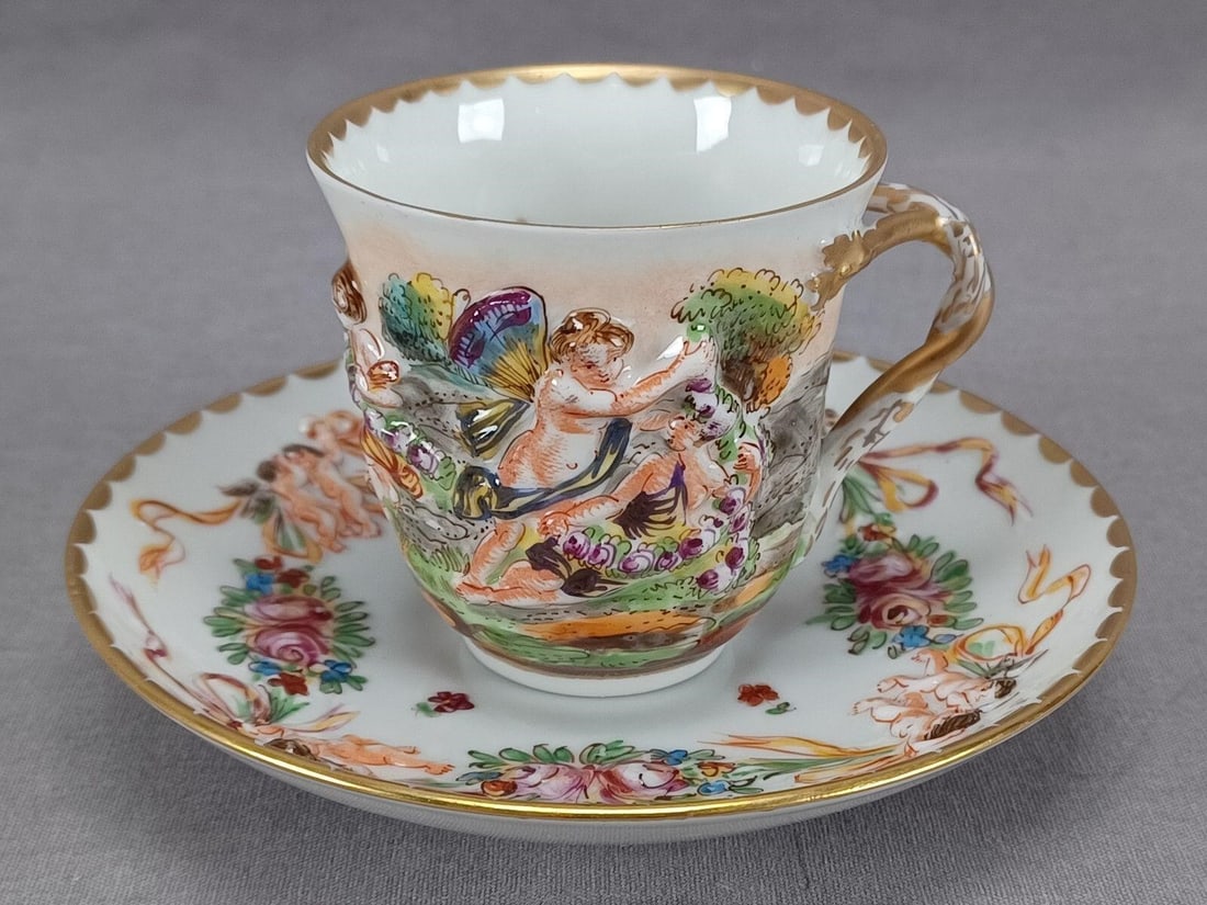 Antique Capodimonte Style Demitasse Cup & Saucer A: Nice antique late 19th century German Capodimonte style porcelain cup and saucer. With relief molded and hand painted Neoclassical scenes on the cup and cherubs with rose swags on the saucer. The cup