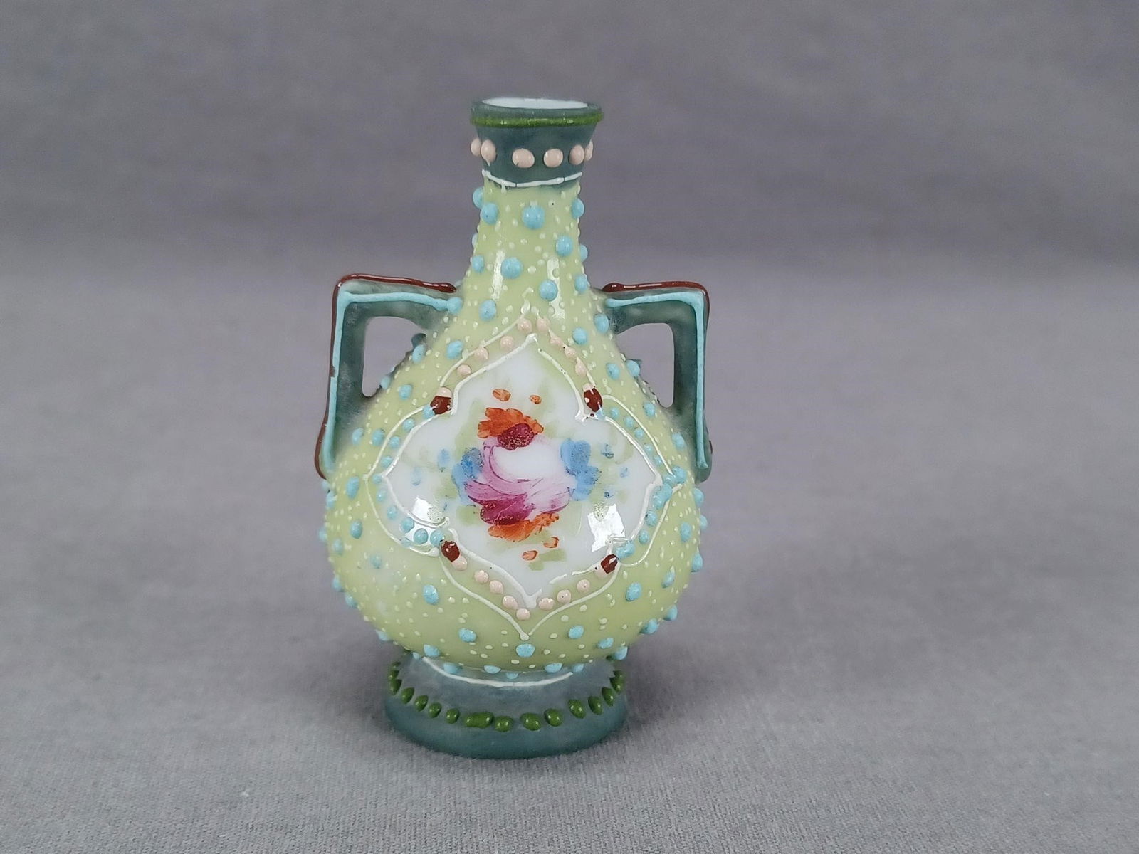 Nippon Raised Enamel Miniature 2 3/4" Green Vase: Nice antique Japanese porcelain vase. Decorated with hand painted pink, blue and red flowers over a white quatrefoil framed by white enamel and pink, red, and turquoise enamel beads. The vase has a mo