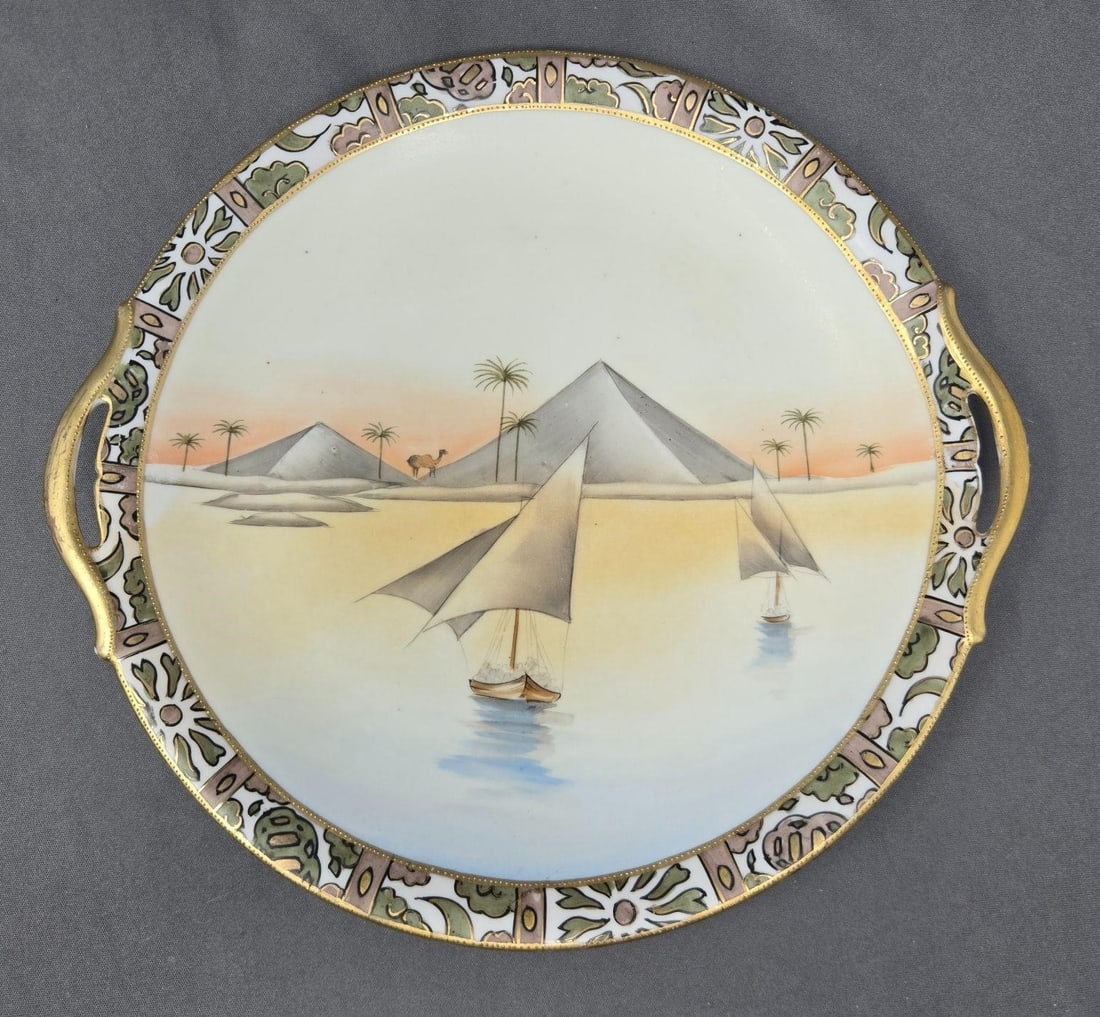Hand Painted Nippon Handled Plate Egyptian Revival: Hand Painted Nippon Egyptian revival handled plate. Design shows pyramids, camel, sailboats and palm trees. Beading on both sides of border. Measures approximately 10 1/2 inches with handles and 9 3/4