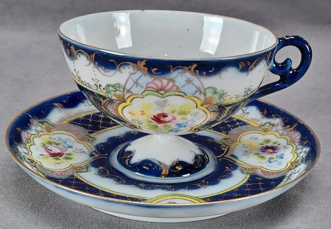 Nippon Hand Painted Floral Cobalt Tea Cup & Saucer: Antique Japanese porcelain pedestal tea cup and saucer. Decorated with hand painted flowers in pink, purple, blue, and yellow framed by yellow and gold scrollwork alternating with gold medallions fram