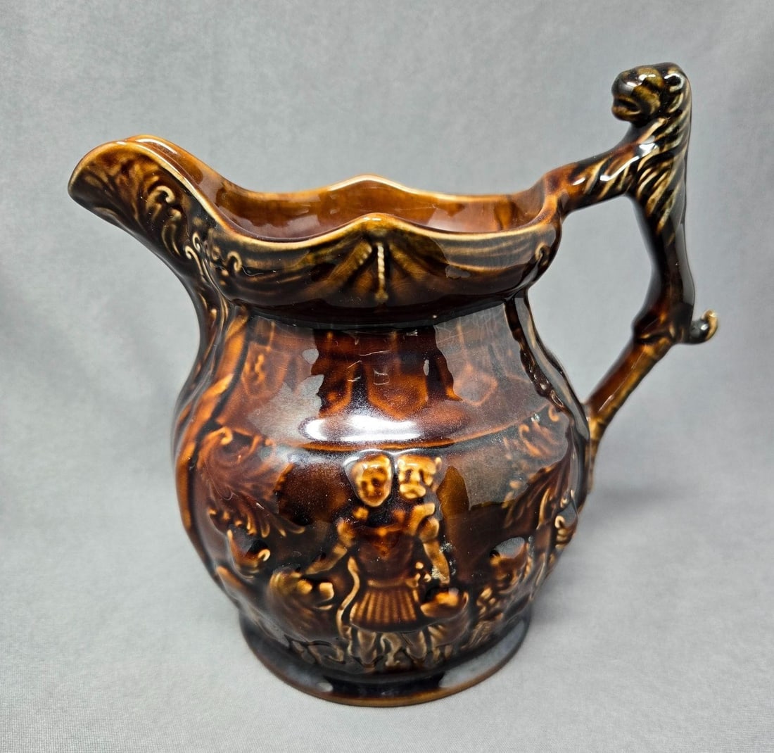 Arthur Wood Rockingham Lions Pitcher c. 1920: Arthur Wood Rockingham pitcher. The design is a man with lions, one is on his back and his hand is on another. The handle is also a lion. Measures approximately 6 3/8 inches tall and 7 inches from spo