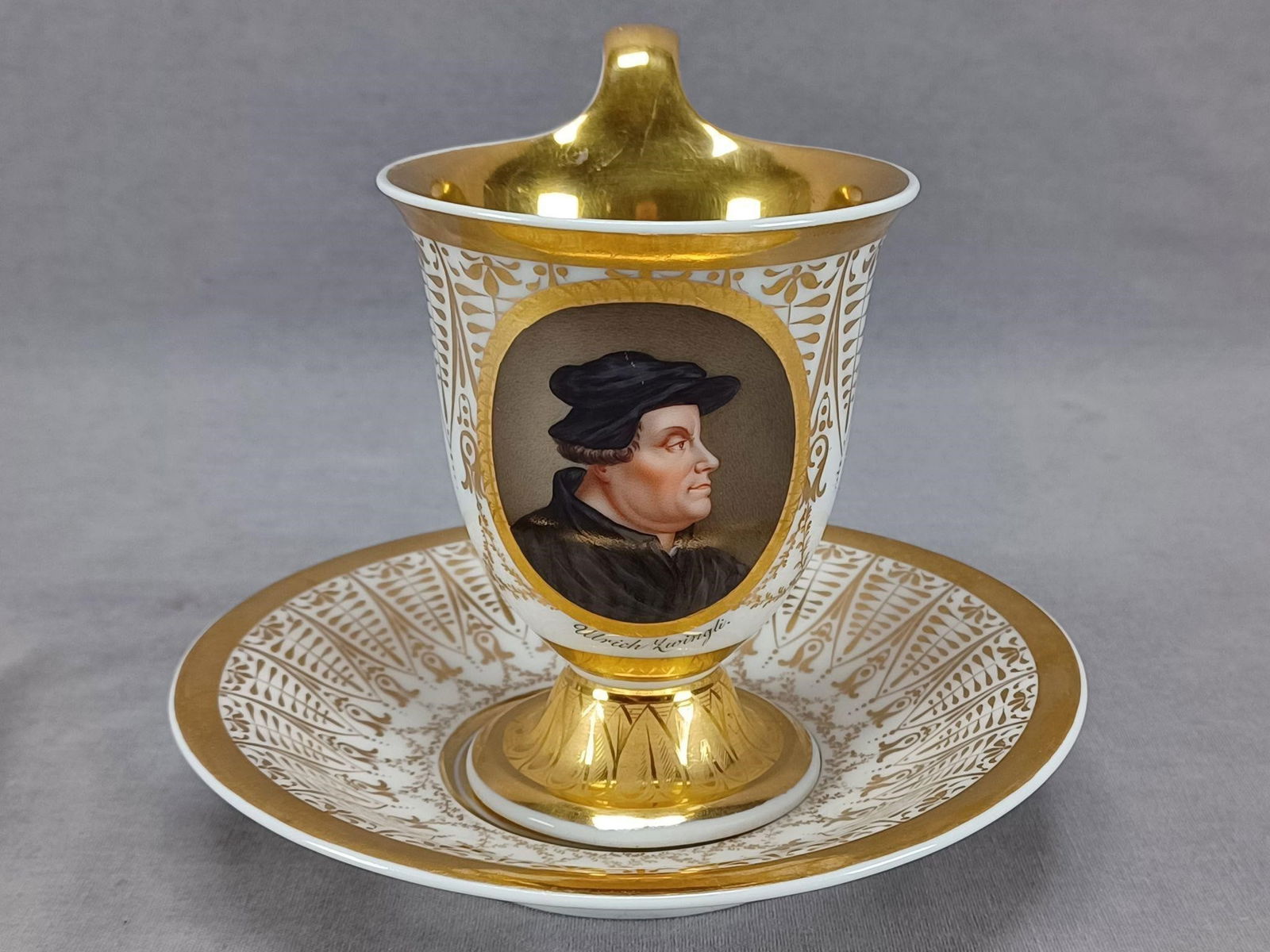KPM Berlin HP Ulrich Zwingli Portrait Cup & Saucer: Antique German porcelain footed empire form cup and saucer. Has a hand painted portrait of Ulrich Zwingli framed in gold gilding. With gold leaves, flowers, and leaf swags. The saucer has a gold