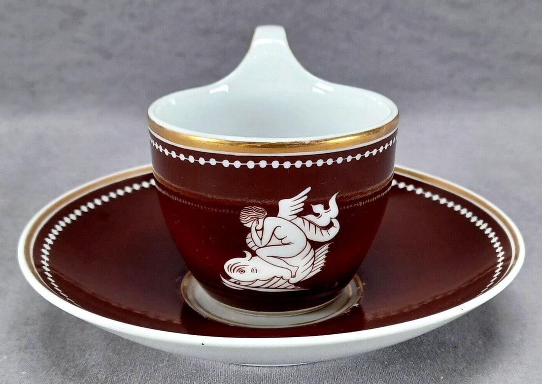KPM Berlin Cherub Riding Dolphin Cup & Saucer: Nice antique mid 19th century German porcelain empire form cup and saucer. The cup has a white cherub riding a Neoclassical cherub opposite the handle. The cup and saucer both have a brown background