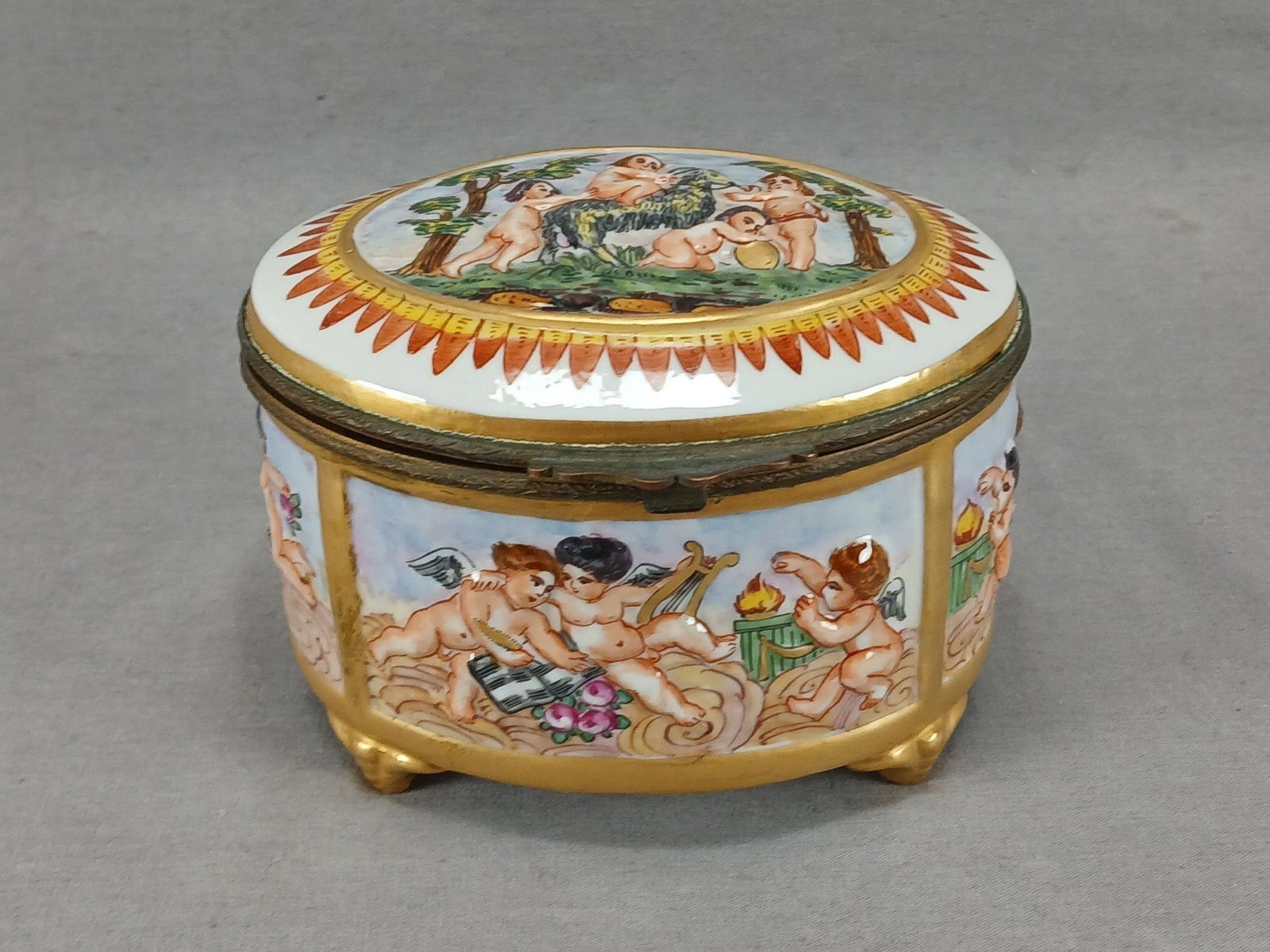 Capodimonte Style Hand Painted Cherubs & Goat Box: Antique European Capodimonte style porcelain box. With relief molded and hand painted cherubs with musical instruments, pink roses, and a goat. With gold gilding around each scene. Gilt brass mounts.