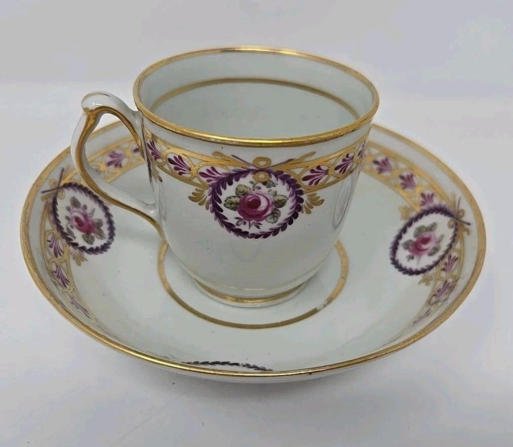 New Hall Cup & Saucer C. 1782-87 Pink Roses: Unmarked New Hall cup and saucer set. Decorated with pink roses, purple laurel wreaths, and a gold and purple band. There are no chips or crazing. There is a star crack on the bottom of the saucer. Th