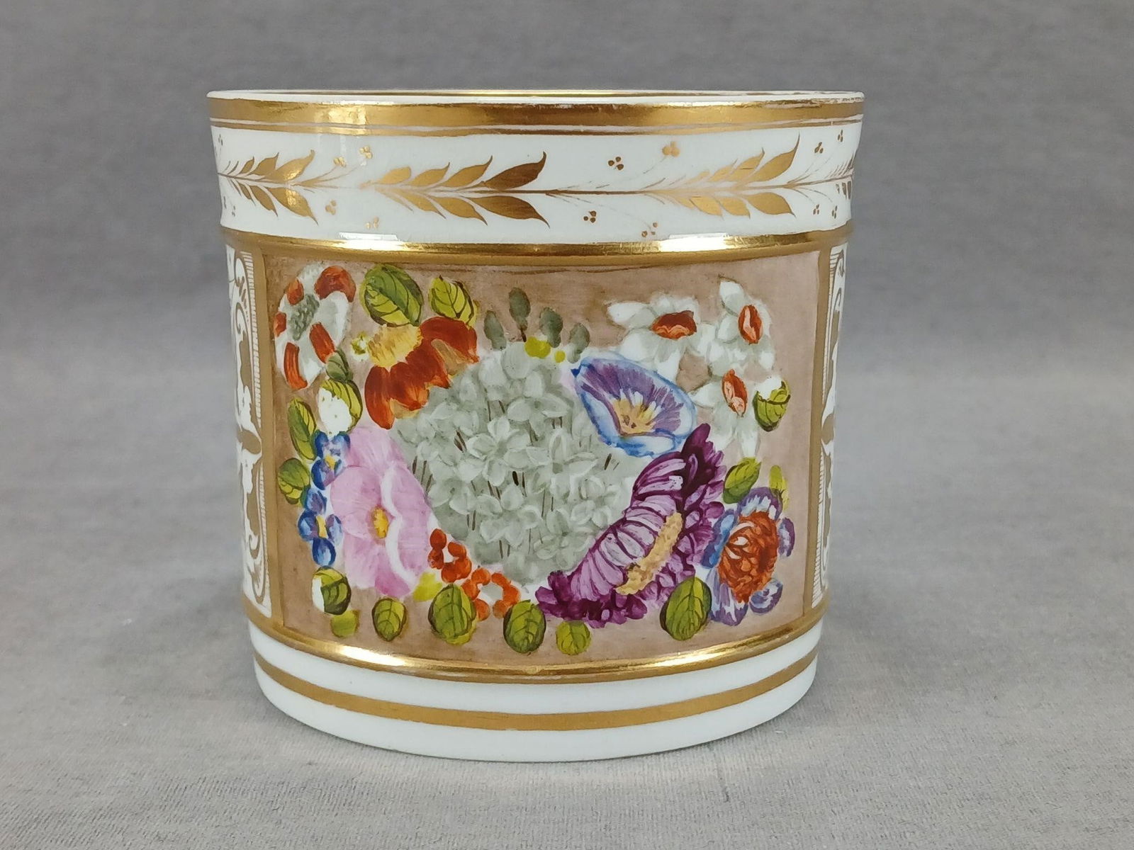 Derby Hand Painted Flowers Mug Circa 1806-1825: Antique British bone china mug. Decorated with hand painted flowers in white, purple, blue, pink, red, and yellow on the front over beige. Has gold gilt leaves around the top and sides. Gold leaves to