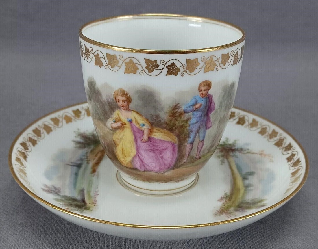 Sevres Hand Painted Watteau Coffee Cup & Saucer: Antique French porcelain coffee cup and saucer. Decorated with a hand painted Watteau scene on the cup. The saucer has hand painted landscape scenes on either side. Gold ivy leaf band around the top o