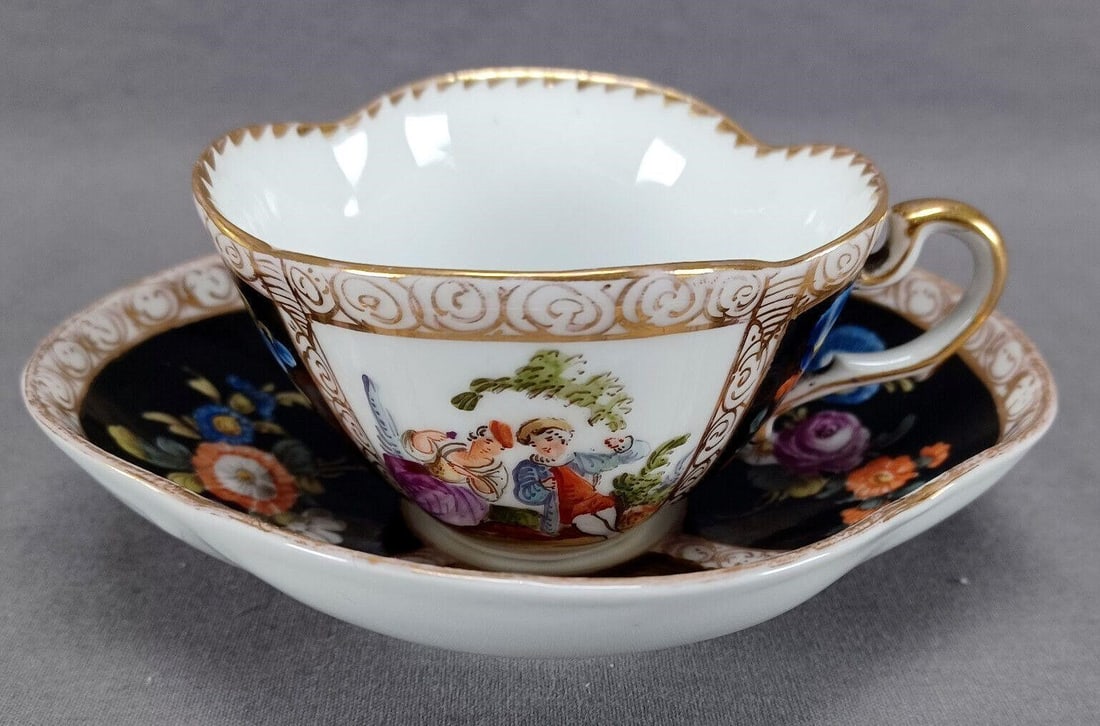 Dresden Watteau Scene Black Tea Cup & Saucer B: Nice antique German porcelain quatrefoil shaped tea cup and saucer. Decorated in alternating panels with hand painted Dresden flowers over black and scenes with hand painted Watteau scenes / courting