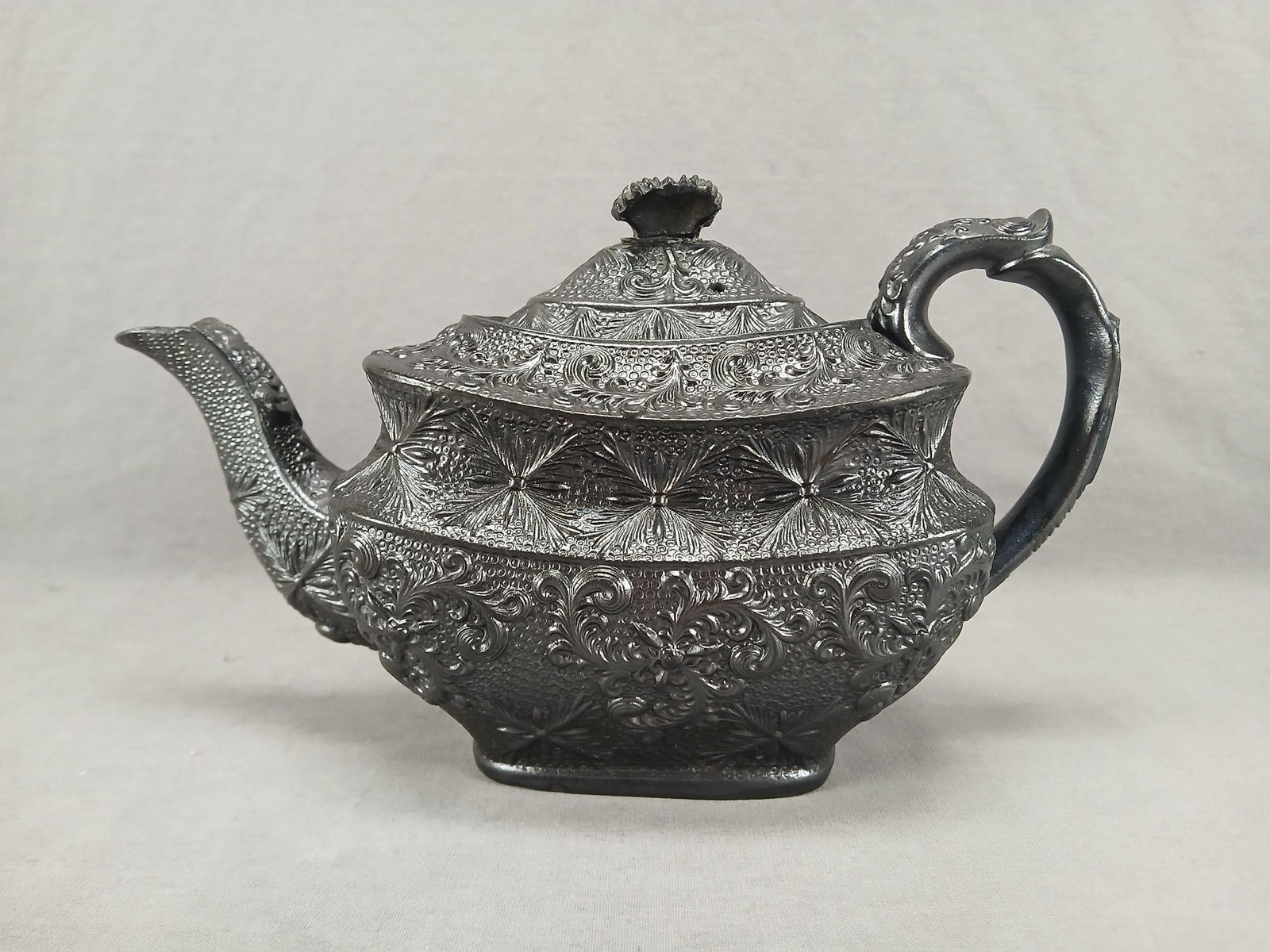 Black Basalt Floral Scrollwork Teapot Circa 1820s: Antique British black basalt teapot. With relief molded floral scrollwork designs. Unmarked. Circa 1820s.Has a chip to the side of the spout, a chip to the rim of the pot, a chip to the foot rim of th