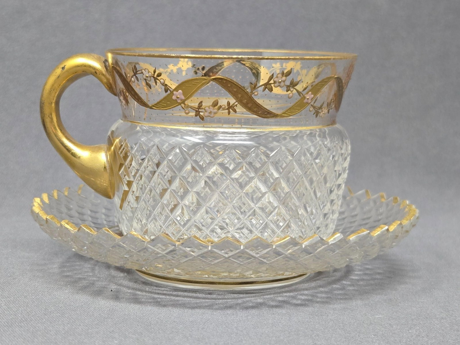 Bohemian Diamond Pattern Cut Glass Cup and Saucer: Bohemian diamond pattern cut glass cup and saucer set. It has an enameled floral garland with a raised gold ribbon design and attached glass handle and a star cut on the base. No chips or cracks. The 