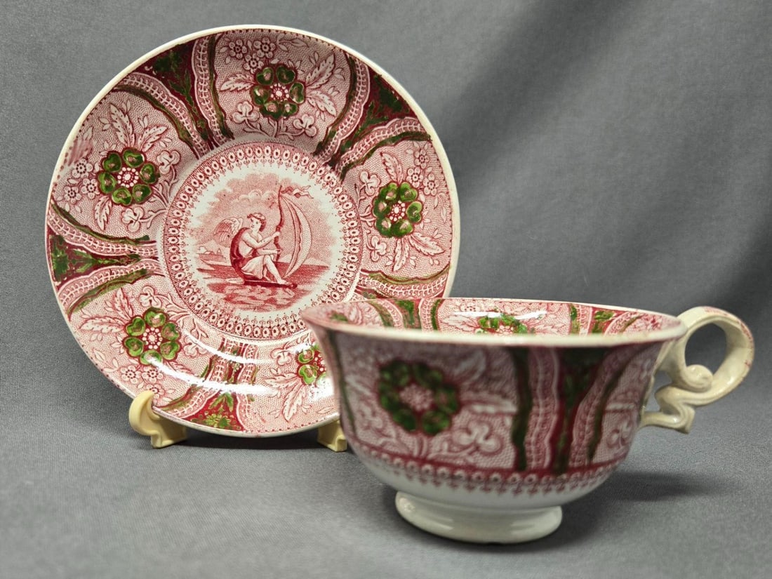 Cupid Pattern Transferware Cup and Saucer: Red transferware hand colored with green accents Cupid pattern # 10865. Earthenware with no chips or cracks. There is crazing which is common with this material. Approximate measurements: The saucer
