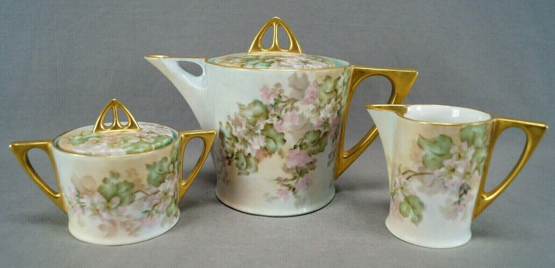 Hutschenreuther Hand Painted Tea Trio: Porcelain tea trio including teapot, creamer, and sugar in an Art Deco shape. Each piece is decorated with hand painted pink and white flowers. With gold gilding along the rim and to handles, spouts, 