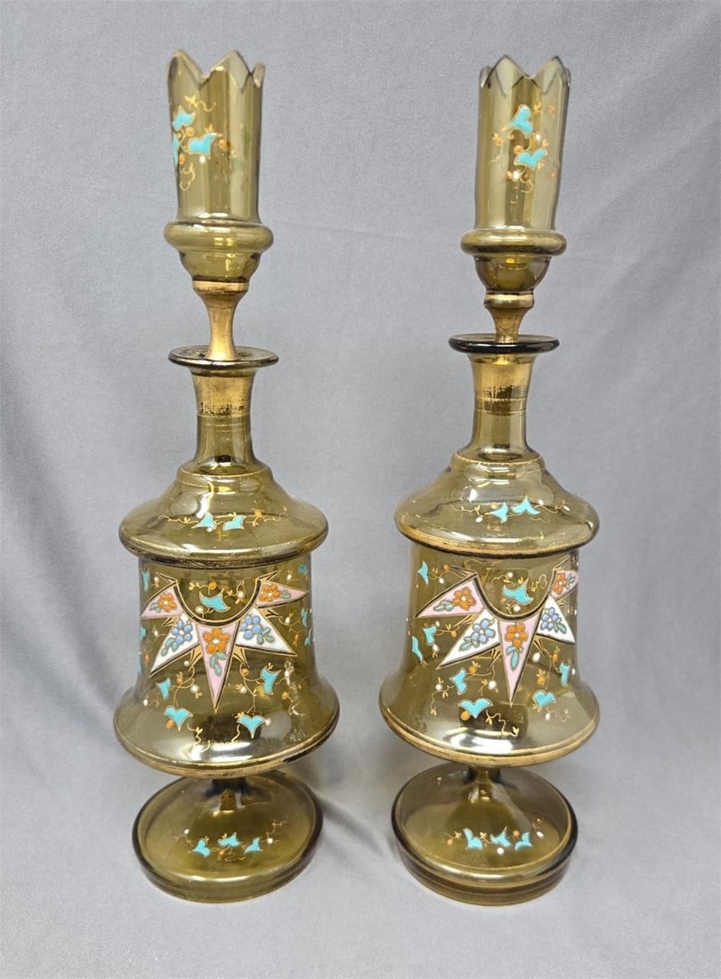 Pair Bohemian Decanters with Shot Glass Stoppers: Pair unusual Bohemian decanters with shot gasses in the stoppers. A greenish amber color with hand enameled design and gold gilt trim. They measure approximately 12' and 12 1/4" as one is a bit taller