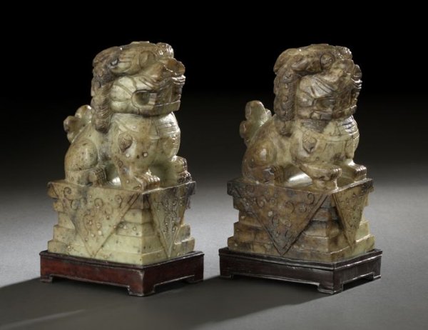 Pair of Chinese Serpentine Figures of Foo Dogs: Pair of Chinese Carved Serpentine Figures of Foo Dogs, each carved of mottled green and russet stone depicting a figure of a Foo dog seated on its haunches atop a plinth base, presented on carved wood