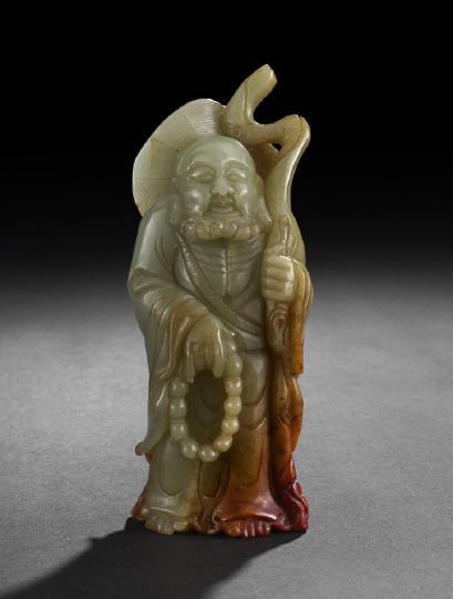 Chinese Carved Jade Figure of a Buddhist Immortal: Chinese Carved Jade Figure of a Buddhist Immortal, 19th/20th century, the celadon stone with russet inclusions and carved as a standing robed and bearded figure with a straw peasant's hat, holding a s