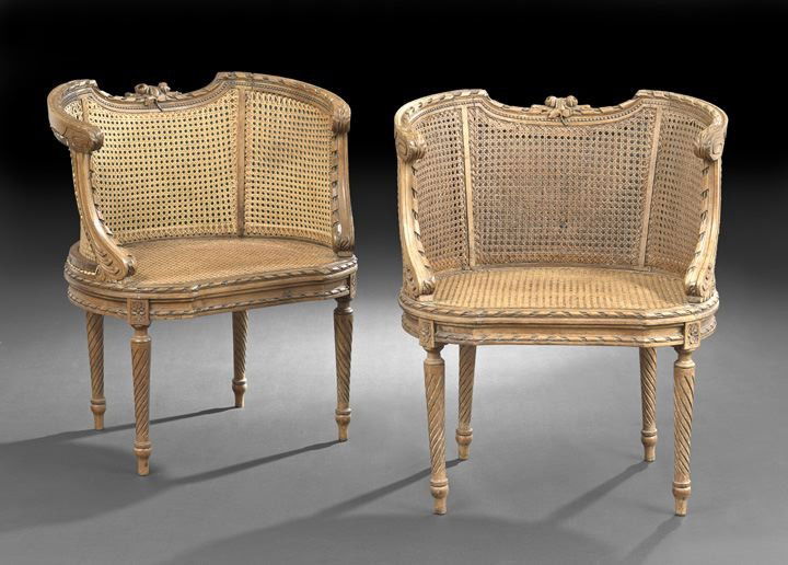 Pair of Louis XVI-Style Fruitwood Bergeres: Pair of Louis XVI-Style Fruitwood Bergeres, early 20th century, each with a low concave caned back surmounted by a floral-and-ribbon-carved crest, joined by caned sides and acanthine arms, the cushion