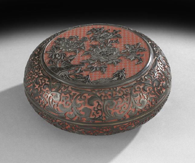 Chinese Two-Color Lacquer Storage Box: Chinese Two-Color Lacquer Storage Box, 20th century, the circular box with fitted dome cover carved in black over cinnabar lacquer with a central scene of peonies and rockwork, the sides carved as scr