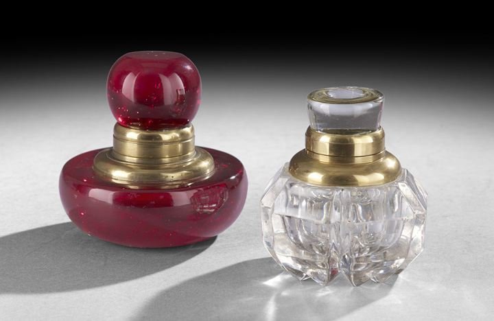 Two Victorian Glass and Brass Inkwells: Two Victorian Glass and Brass Inkwells, fourth quarter 19th century, English, the ruby glass example with a bun-shaped base and a pontil mark, the globular cap with a polished top attached with a heav