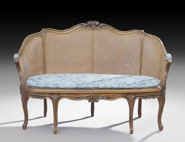 Provincial Louis XV-Style Fruitwood Settee: Provincial Louis XV-Style Fruitwood Settee, 19th century, the domed and carved back surmounted by a floral crest, joined by downswept arms to the cushioned caned seat above a shaped floral-carved apro